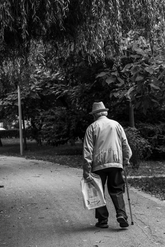 This is a black and white image of an elderly man walking with a can in a par. The man is carrying a package.