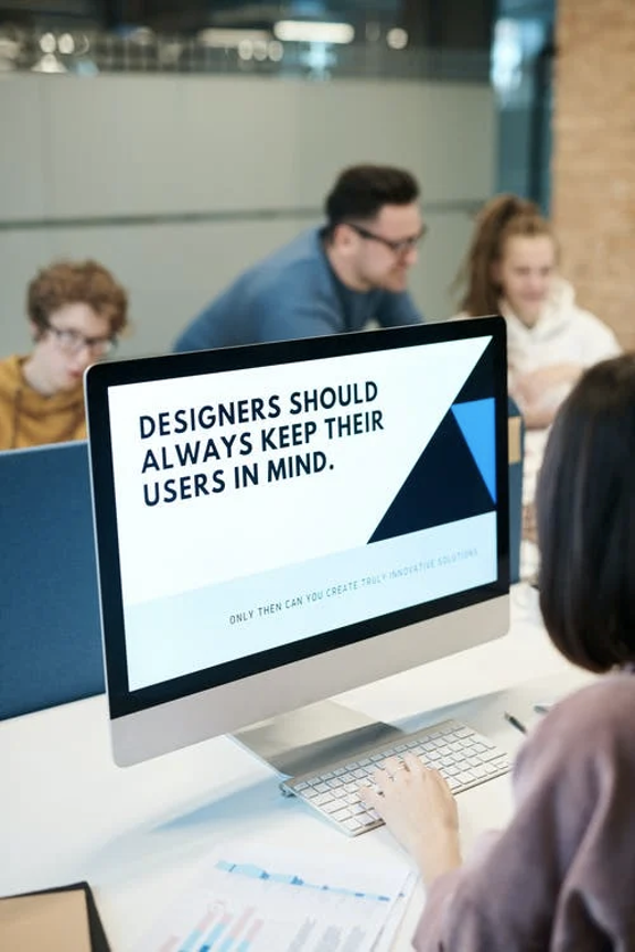 This is a photo of a woman working on a computer. The screen reads "DESIGNERS SHOULD ALWAYS KEEP THEIR USERS IN MIND."